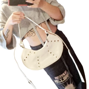 Vit Y2K handväska med platta nitar från tidigt 2000-tal  - Underbar vit Y2K handväska med platta nitar från tidigt 2000-tal. Väldigt välbevarad, fräsch insida, näst intill nyskick. 30 cm bred. Ställbara band så den kan även bäras som axelremsväska. Använd gärna köp-knappen 💖💖 & följ min profil för fler nyheter! 🥰