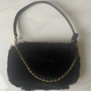 Pälsväska - Väska i pälsimitatipn från Guess. Axelband och dekorativ kedja. Väskan är i nyskick då den endast använts en gång.