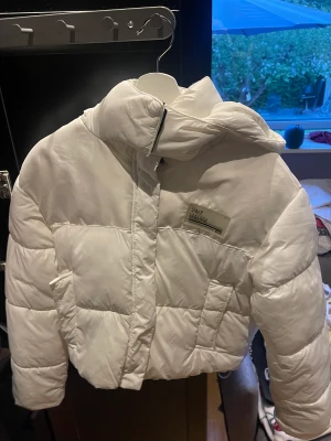 Puffer jacket - Vit croppad pufferjacka med luva från Bershka. Använd under kort period men kommer tyvärr inte till användning längre. Har 2 fickor fram och en innerficka. Går även att justera långst ner på jackan. Har både dragkedja och knappar!