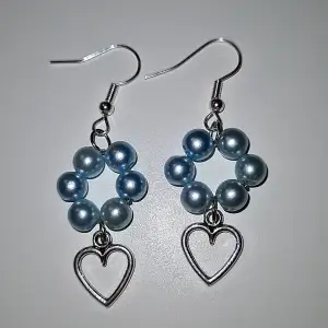 Ett par handgjorda blå-blomformade örhängen med ett varsitt hängande hjärta nedtill. Ej äkta silver så kan innehålla nickel. 