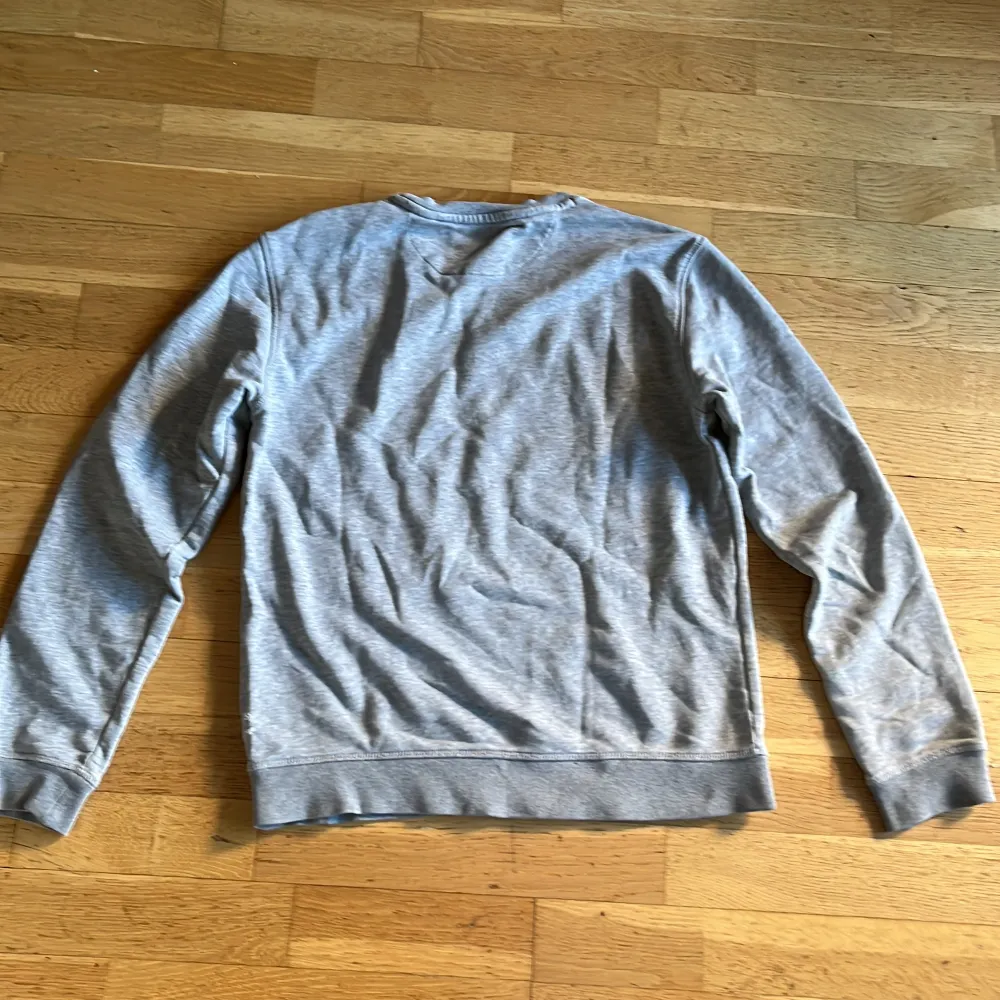 Säljer en stilren grå sweatshirt från GANT med broderad logga på bröstet. Tröjan har rund hals och långa ärmar, perfekt för en avslappnad look. Den är tillverkad i mjukt material för extra komfort. Den har dessvärre två små hål vid sidan.. Neuletakit & Villapaidat.