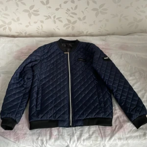 Blå quiltad bomberjacka från Simelp - Snygg blå quiltad bomberjacka från Simelp med dragkedja framtill. Jackan har ribbade muddar vid ärmslut och nederkant samt en cool patch med texten 'BEST FASHION' på ärmen. Perfekt för en stilren look.