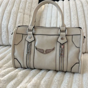 Beige handväska med vingdetalj - Jätte fin beige handväska från Zadig köpt begagnat ägaren sa att hon inte viste om den va äkta ädå hon inte hade med något kvitto elr box aldrig använt då jag använder guld smycken och de är silver detalje storlek 25/16