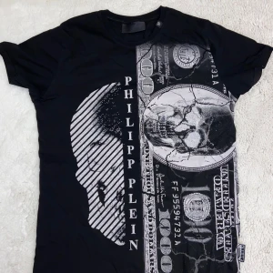 Svart t-shirt från Philipp Plein. Den passar också XL och L på sättet man stylar den. - Cool svart t-shirt från Philipp Plein med ett unikt tryck av en dödskalle och sedlar på framsidan. Perfekt för dig som vill sticka ut med en edgy stil. T-shirten har korta ärmar och en normal passform.