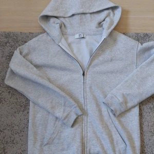Grå hoodie från bvaldi - Säljer en stilren grå hoodie från Valdi med dragkedja och glittrig logga på bröstet. man får även en tag man kan ta på tröjan gratis HELT NY ALDRIG ANVÄNT.