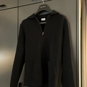 Svart cardigan från John Henric - Säljer en skön cardigan från John Henric med dragkedja framtill. Tröjan har långa ärmar och en klassisk krage. Perfekt för en elegant och avslappnad look.