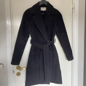 Svart kappa med bälte - Svart kappa från VILA i storlek XS. Den har knytbart bälte i midjan, knappar samt två öppna fickor.