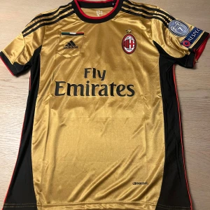 Guld fotbollströja från Adidas - Snygg guld fotbollströja från Adidas med AC Milan-emblem och Fly Emirates-tryck. Tröjan har korta ärmar och svarta detaljer på sidorna. Champions League-märke på ärmen och spelarnamn och nummer 'KAKA 22' på ryggen.
