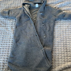 Åkpåse - Mysig grå stickad babyoverall med huva och knappar framtill. Perfekt för att hålla de små varma under kyliga dagar. Overallen har en omlott-design och är ribbad vid kanterna för extra komfort.
