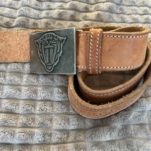 TIGER OF SWEDEN BÄLTE - Snyggt brunt läderbälte med ett unikt metallspänne som har ett djurmotiv. Bältet har vita sömmar som ger en fin kontrast och en rustik känsla. Perfekt för att ge en extra touch till din outfit.