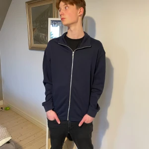 Mörkblå tröja med dragkedja - Säljer en stilren mörkblå tröja med dragkedja. Perfekt för en avslappnad look. Tröjan har långa ärmar och en hög krage som kan bäras uppvikt eller nedvikt. Passar bra till jeans eller chinos. Kommer från 157