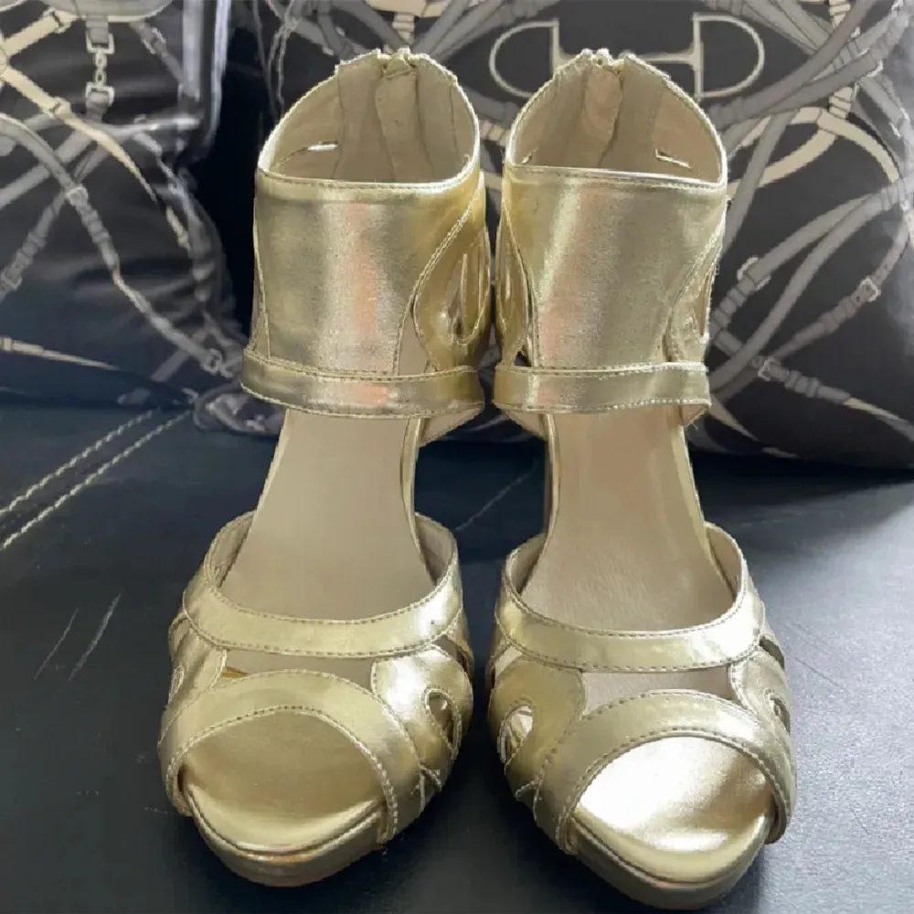 Snygga guldiga klackskor med unika utskärningar och öppen tå. Perfekta för att ge en glamorös touch till din outfit. Skorna har en elegant design med hög klack och dragkedja baktill för enkel på- och avtagning. Oanvända. . Kengät.