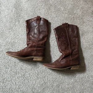 Bruna cowboy boots från Zara - Säljer ett par snygga bruna cowboy boots från Zara. De är tillverkade i skinn med dekorativa broderier och har en spetsig tå. Perfekta för en cool och avslappnad stil.