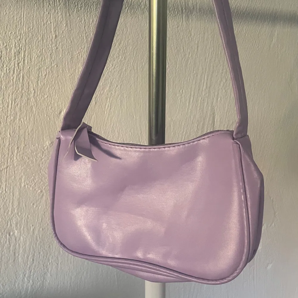 Snygg lila axelväska i syntetmaterial med en söt rosett på sidan. Väskan har en praktisk dragkedja för säker förvaring av dina saker. Perfekt för att ge en färgklick till din outfit!. Laukut & Käsilaukut.