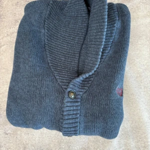 Blå kofta från Chaps - Säljer en stilren blå kofta från Chaps i 100% bomull. Koftan har en ribbad krage och knappar framtill. Perfekt för en avslappnad och bekväm stil.