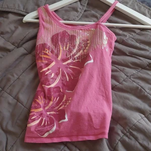 Y2k linne - Säljer ett rosa linne med stora blommor i lila och guldiga detaljer. Linnet har smala axelband och är perfekt för sommardagar. Detaljerna ger en glittrig effekt som gör det extra speciellt.