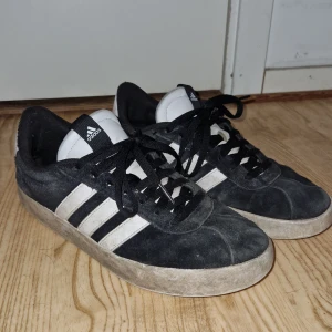 Svarta sneakers från Adidas - Säljer ett par klassiska svarta sneakers från Adidas med vita ränder och snörning. Skorna har en vit sula och är perfekta för vardagsbruk. De ikoniska tre ränderna ger en tidlös look.