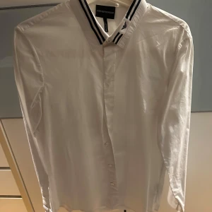 Vit skjorta från Emporio Armani - Snygg vit skjorta från Emporio Armani med stilrena svarta ränder på kragen. Skjortan har en slim passform och är perfekt för en elegant look. Den är långärmad och har knappar framtill.