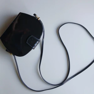 Svart axelväska  - Snygg svart axelväska från Giovanni Couture med en elegant och glansig finish. Väskan har en smal axelrem och guldfärgade detaljer som ger en lyxig touch. Perfekt för att bära dina nödvändigheter med stil.