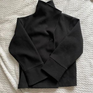 Svart kappa med hög krage - Snygg svart kappa med hög krage och knappar framtill. Perfekt för kyligare dagar med sin stilrena design och långa ärmar. En tidlös klassiker som passar till många tillfällen.