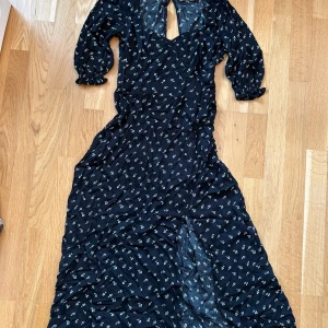 Svart blommig långklänning - Somrig svart långklänning med små vita blommor. Klänningen har trekvartsärmar och en v-ringad design med en liten öppning i ryggen. Perfekt för en elegant, somrig look.