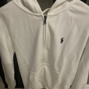 Vit hoodie från Ralph Lauren - Säljer en stilren vit hoodie från Ralph Lauren med dragkedja och det klassiska logotypbroderiet på bröstet. Perfekt för en avslappnad look. Hoodien har långa ärmar och en bekväm passform.