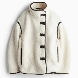 Vit teddyjacka med svarta detaljer - Säljer en mysig vit teddyjacka med svarta kantdetaljer och knäppning framtill från hm. Jackan har en hög krage och långa ärmar, perfekt för kyliga dagar. Den är både stilren och bekväm. Slutsåld på hemsidan och jätte populär!💕