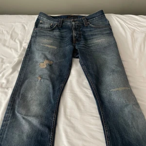 Blå jeans från Nudie Jeans - Snygga blå jeans från Nudie Jeans med slitna detaljer och en klassisk femficksdesign. Jeansen har en knappgylf och är tillverkade i denim med en cool, tvättad look. Perfekta för en avslappnad stil.