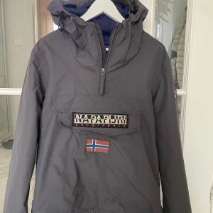 Napapijri vindjacka - Säljer en grå vindjacka från Napapijri med huva och dragkedja. Jackan har en stor ficka framtill med logotyp och en norsk flagga. Perfekt för blåsiga dagar.