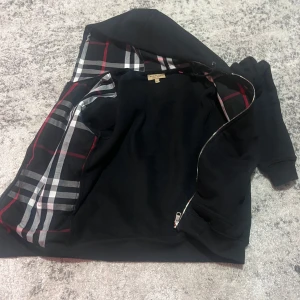 Burberry kofta - Snygg svart huvtröja med dragkedja och ett stilrent rutigt foder i grått, vitt och rött. Perfekt för en casual look med en touch av elegans. Huvan har justerbara snören och tröjan har långa ärmar.