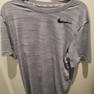 Grå t-shirt från Nike - Säljer en grå t-shirt från Nike med ett diskret mönster och den klassiska Nike-loggan i svart på bröstet. T-shirten har en rund hals och korta ärmar, perfekt för träning eller vardagsbruk.