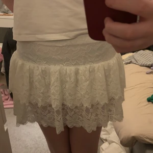 Vit spetskjol med volang - Säljer en söt vit kortkjol i spets med volangdetaljer. Kjolen har en elastisk midja och har vita shorts under. 