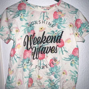 Blommig t-shirt från KappAhl - Säljer en somrig t-shirt från KappAhl med färgglatt blommönster och texten 'Weekend Waves' på framsidan. Perfekt för soliga dagar men även som en tröja att kunna ha på sig utomlands eller en dag till typ stranden! 🌸