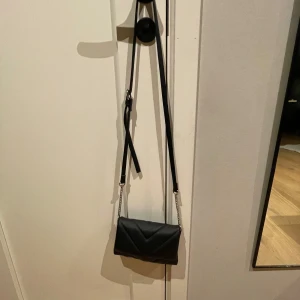 Svart quiltad axelväska med kedjedetaljer - Snygg svart axelväska med quiltad design och kedjedetaljer. Väskan har en justerbar axelrem och en stilren look som passar perfekt för olika tillfällen. Den är kompakt men rymmer det viktigaste du behöver ha med dig.