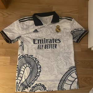 Real Madrid tröja  - Säljer en vit fotbollströja från Adidas med ett coolt drakmönster i svart. Tröjan har korta ärmar och en svart krage. Real Madrids emblem är broderat på bröstet. Perfekt för fotbollsfans som vill sticka ut!