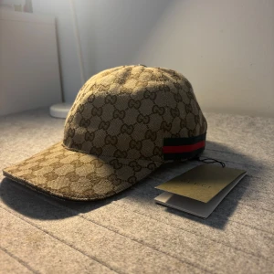 Beige gucci keps - Snygg beige keps från Gucci med det ikoniska monogrammönstret. Kepsen har en justerbar rem baktill och detaljer i rött och grönt på sidan. Perfekt för att ge din outfit en lyxig touch. Storlek ”M”