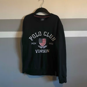 Svart tröja från Vinson Polo Club - Säljer en svart tröja från Vinson Polo Club med ett tryck av en amerikansk flagga och texten 'Polo Club Vinson'. Tröjan har långa ärmar och en rund halsringning. Perfekt för en avslappnad stil.