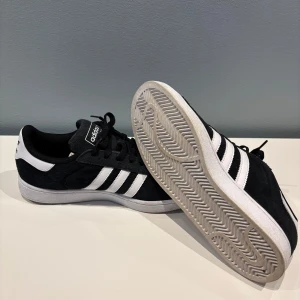 Svarta Adidas Campus 2 sneakers - Säljer ett par klassiska svarta Adidas Campus sneakers med vita detaljer och de ikoniska tre ränderna på sidan. Skorna har snörning och är tillverkade i mocka med en vit sula. Perfekta för en stilren och avslappnad look. I princip helt nya, använt ca 2-3 gånger max. 