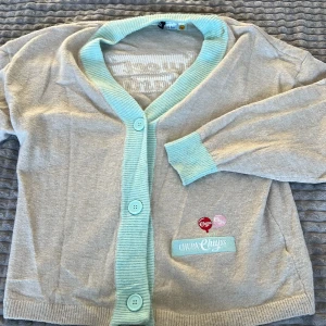 Beige kofta från Chupa Chups - Säljer en beige kofta från Chupa Chups med mintgröna detaljer och knappar. Koftan har ett broderat Chupa Chups-märke och texten 'Sweet 'n tasty' på framsidan. Perfekt för dig som gillar en unik och lekfull stil!