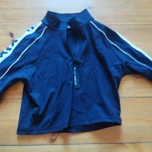 Svart jacka från Elly Pistol - Snygg svart jacka från Elly Pistol med dragkedja framtill och coola vita detaljer på ärmarna. Jackan har en sportig look med tryck på ryggen och axlarna. Perfekt för en trendig och avslappnad stil.