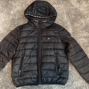 Svart dunjacka med huva - Säljer en svart dunjacka från Tommy Hilfiger med huva och dragkedja. Jackan har en quiltad design och två fickor med dragkedjor framtill. Perfekt för kyliga dagar.