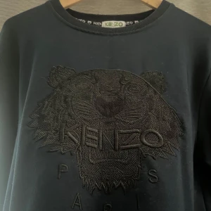 Svart sweatshirt från Kenzo - Strl xxl men sitter som L Snygg svart sweatshirt från Kenzo med ett broderat tigerhuvud och logotyp på framsidan. Tröjan har långa ärmar och en rund halsringning. Perfekt för en stilren look.