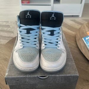 Nike Air sneakers med ljusblå detaljer - Snygga Nike Air sneakers i vitt med ljusblå detaljer och svarta accenter. Skorna har en klassisk hög modell med snörning och den ikoniska Nike-loggan på sidan. Perfekta för en stilren look.