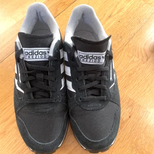 Svarta Adidas sneakers med vita detaljer - Snygga svarta Adidas Treziod 2.0 sneakers med klassiska vita ränder och detaljer. Skorna har snörning och en bekväm passform, perfekt för vardagsbruk. Nästan oanvända. 