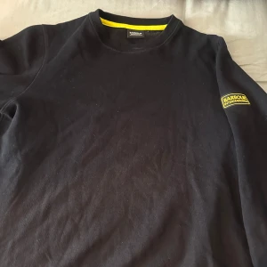 Svart tröja från Barbour - Säljer en svart tröja från Barbour med en gul detalj på insidan av kragen och en logga på ärmen. Tröjan har en klassisk rund hals och långa ärmar. Perfekt för en stilren och avslappnad look. Detta är en L ordinarie men har krympt till M 