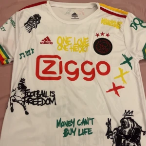 Vit fotbollströja från Adidas - Säljer en vit fotbollströja från Adidas med färgglada tryck och text som 'One Love One Heart' och 'Football is Freedom'. Tröjan har korta ärmar och ett Ajax-märke på bröstet. Perfekt för fotbollsfans som vill sticka ut! Snygg att ha till sommarn🌟🌟