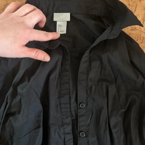 Svart skjorta från H&M - Fin svart skjorta från H&M med klassisk krage och knappar framtill. Perfekt för en stilren look. Skjortan har långa ärmar och är tillverkad i ett lätt material.