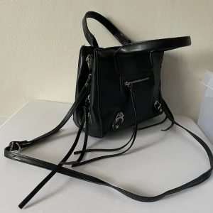 Svart axelremsväska med dragkedja - Svart axelremsväska i skinnimitation med dragkedja och justerbar axelrem. Väskan har två handtag och flera fack på insidan, bland annat ett fack med dragkedja.