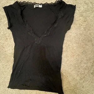 Svart topp med spetsdetaljer från Eve - Säljer en svart topp från Eve med v-ringning och spetsdetaljer runt halsen. Toppen har korta ärmar och en broderad logga längst ner. Perfekt för en stilren look.