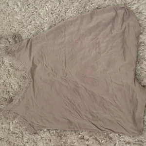 Beige linne med spetsdetaljer - Säljer ett stilrent beige linne med vackra spetsdetaljer längs halsringningen och axelbanden. Perfekt för en avslappnad och chic look. Axelbanden är justerbara för bästa passform. Köpt på Ullared och typ aldrig använt men är ändå lite lite nopprigt på fram och baksida, det är inget man lägger märke till. Kan användas som pyjamas eller till vardagen.